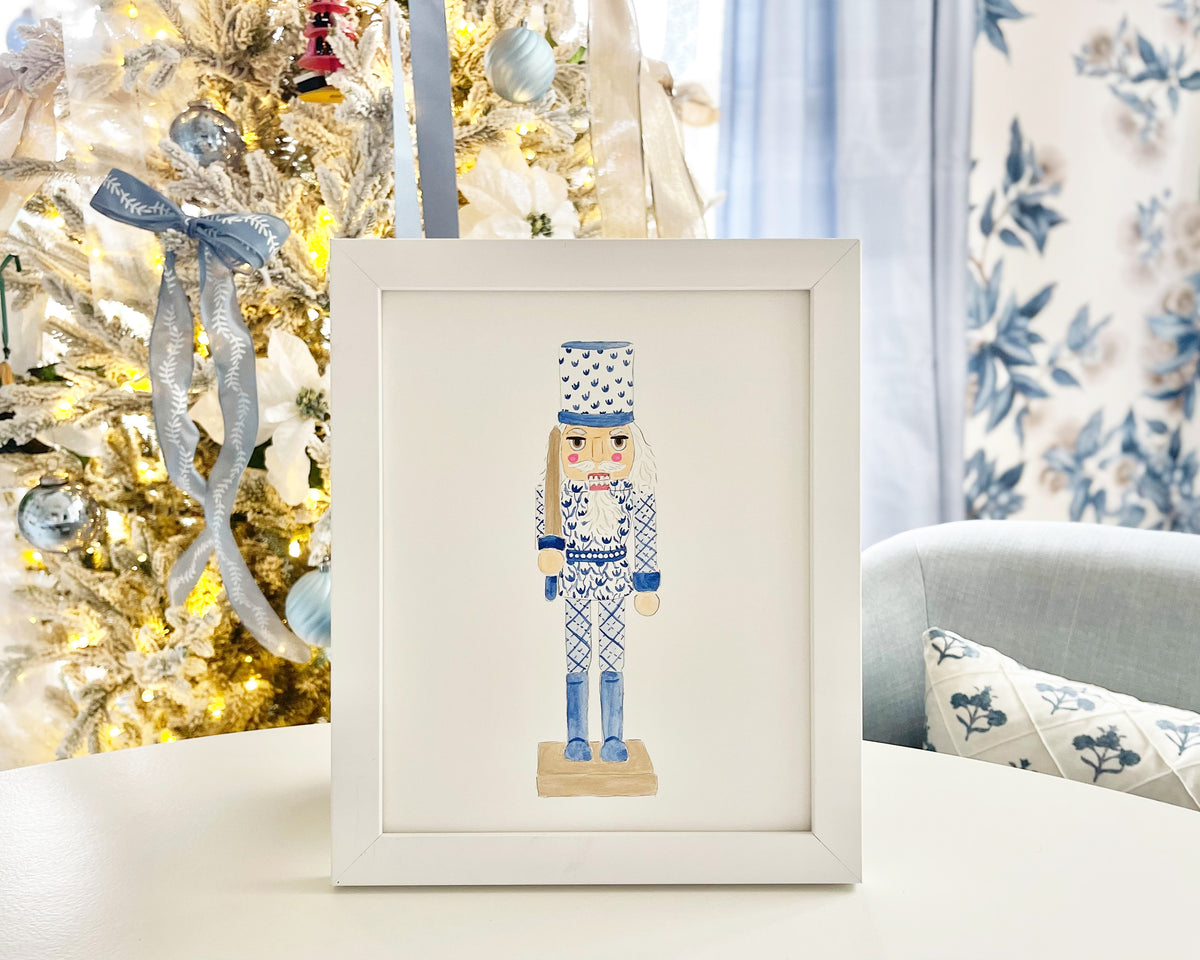 A framed watercolor painting of a nutcracker displayed on a table with a Christmas tree in the background.