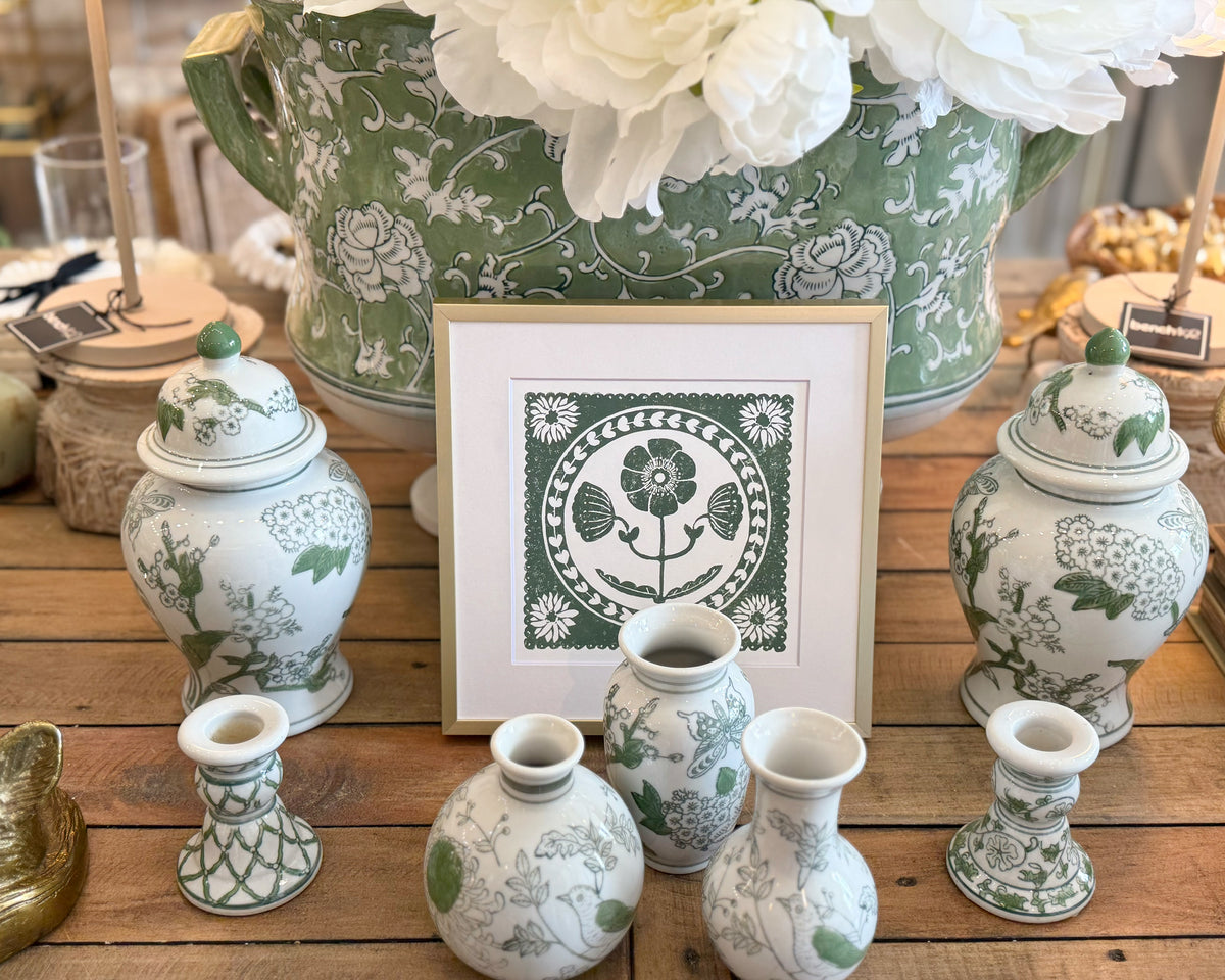 Decorative green and white vases and candles on a wooden surface with a framed print.