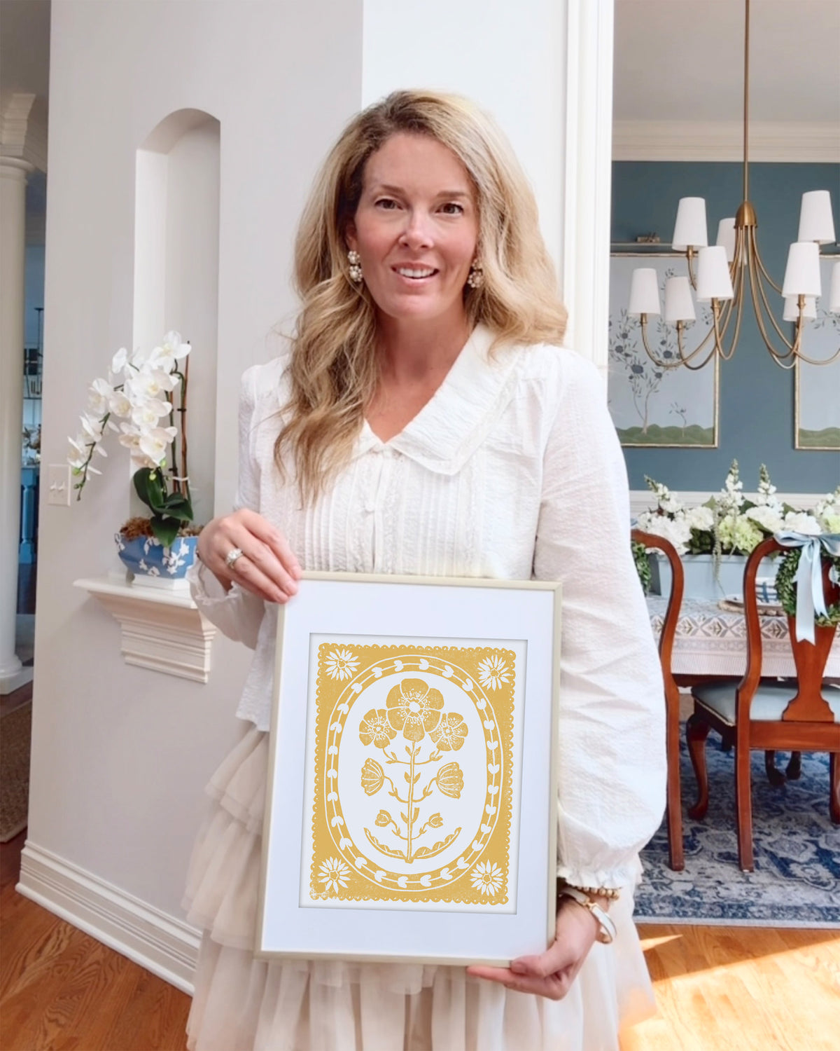 Woman holding a framed mustard yellow and white flower artwork in a home setting