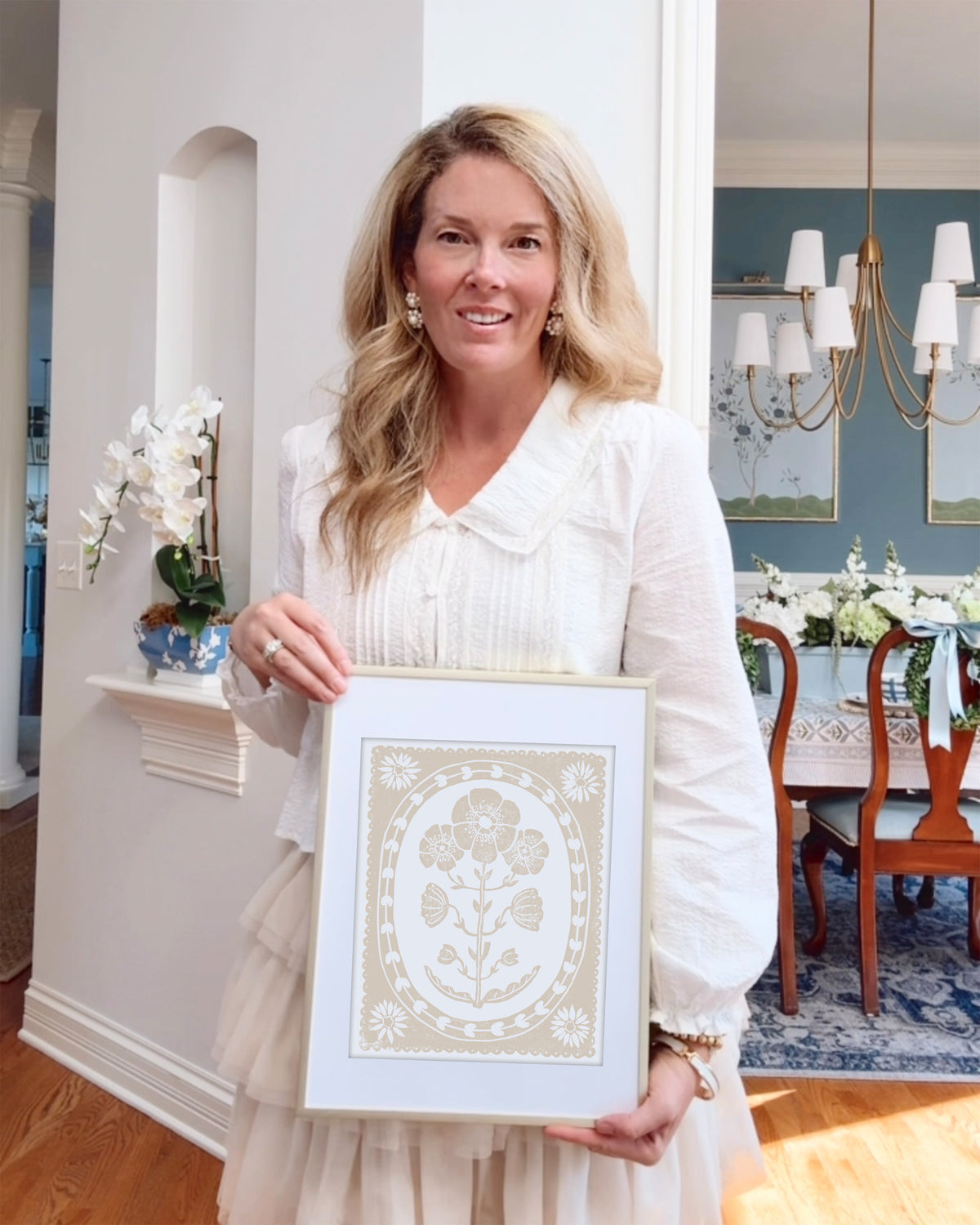 Woman holding a framed beige and white floral artwork in a home setting