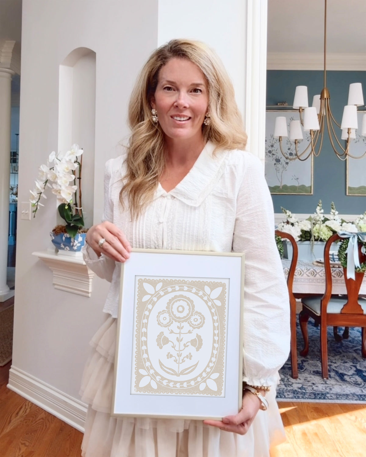 Woman holding a framed beige and white floral artwork in a home setting