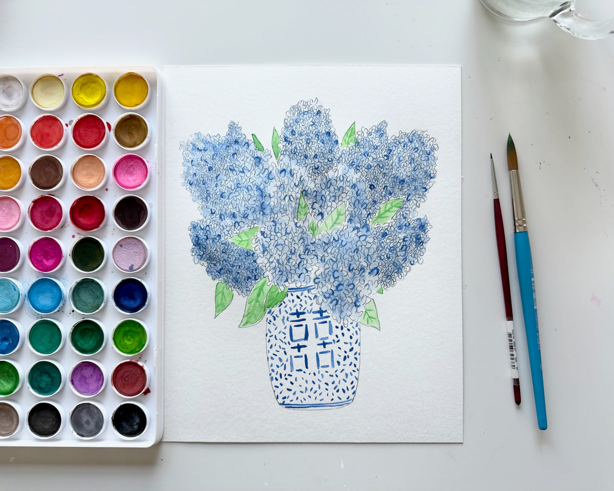 Watercolor painting of blue flowers in a vase on a white paper with a color palette and brushes beside it.