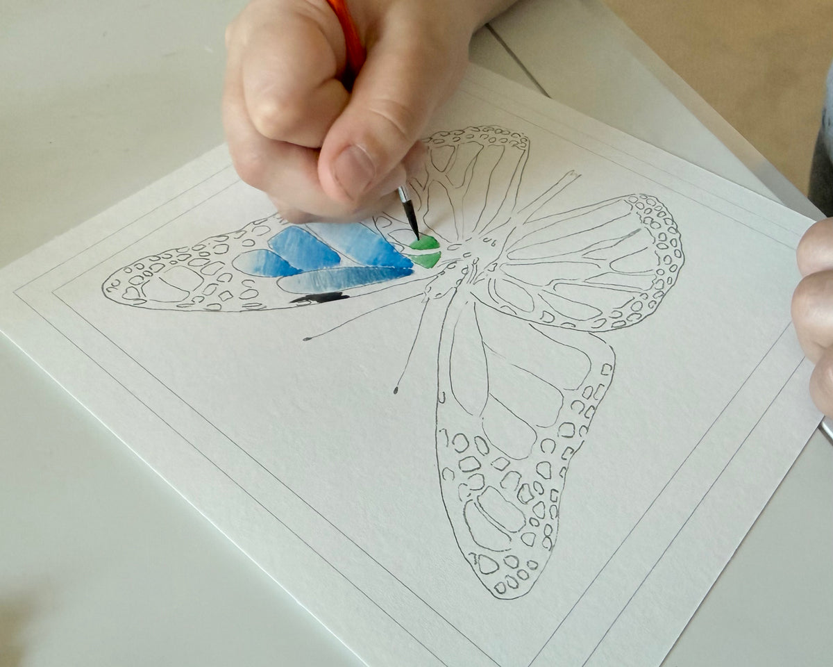 Hand coloring a butterfly illustration with a paintbrush on a piece of paper.