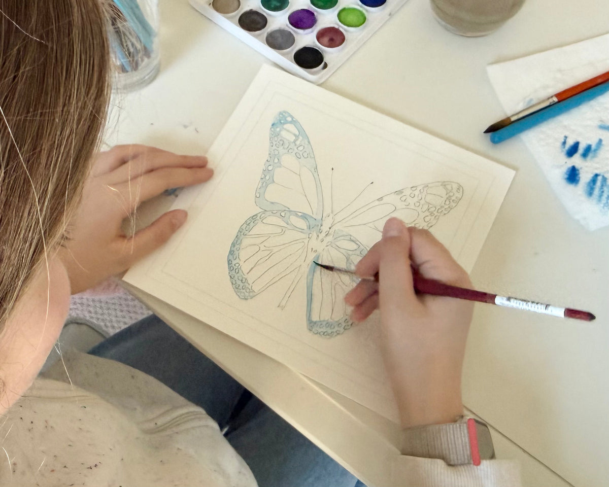 Person painting a butterfly design on a wooden board with watercolors and brushes.