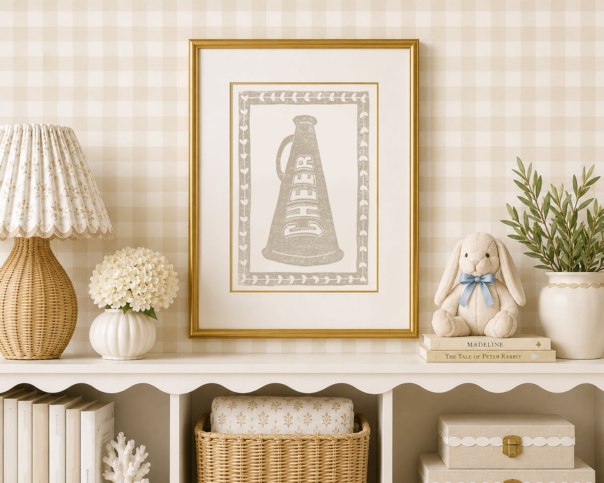 A sand tan and white cheerleading megaphone art print in a gold matted frame on a styled shelf in a tan and white bedroom