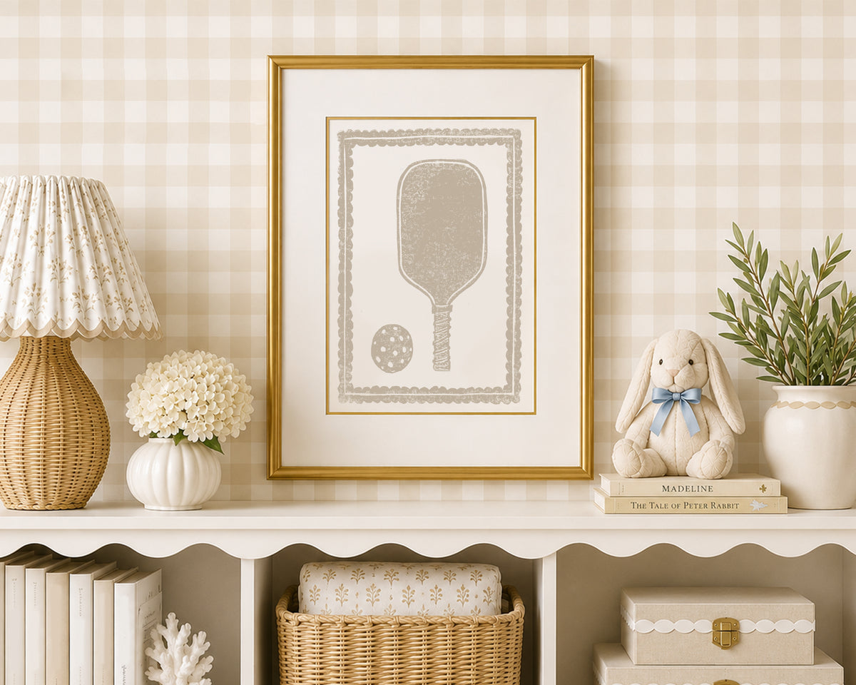 A sand tan and white pickleball racket art print in a gold matted frame on a styled shelf in a tan and white bedroom