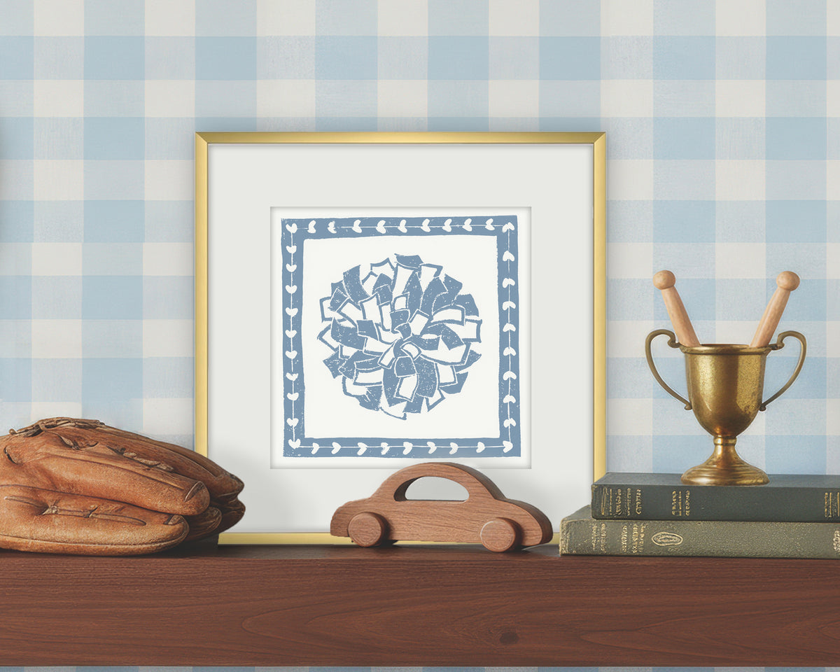 Framed artwork of a blue and white cheerleading pom pon on a white background, displayed on a shelf with a checkered wall.