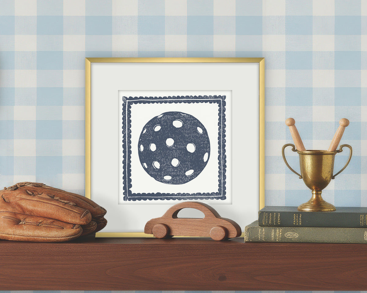 Framed artwork of a blue and white pickleball on a white background, displayed on a shelf with a checkered wall.