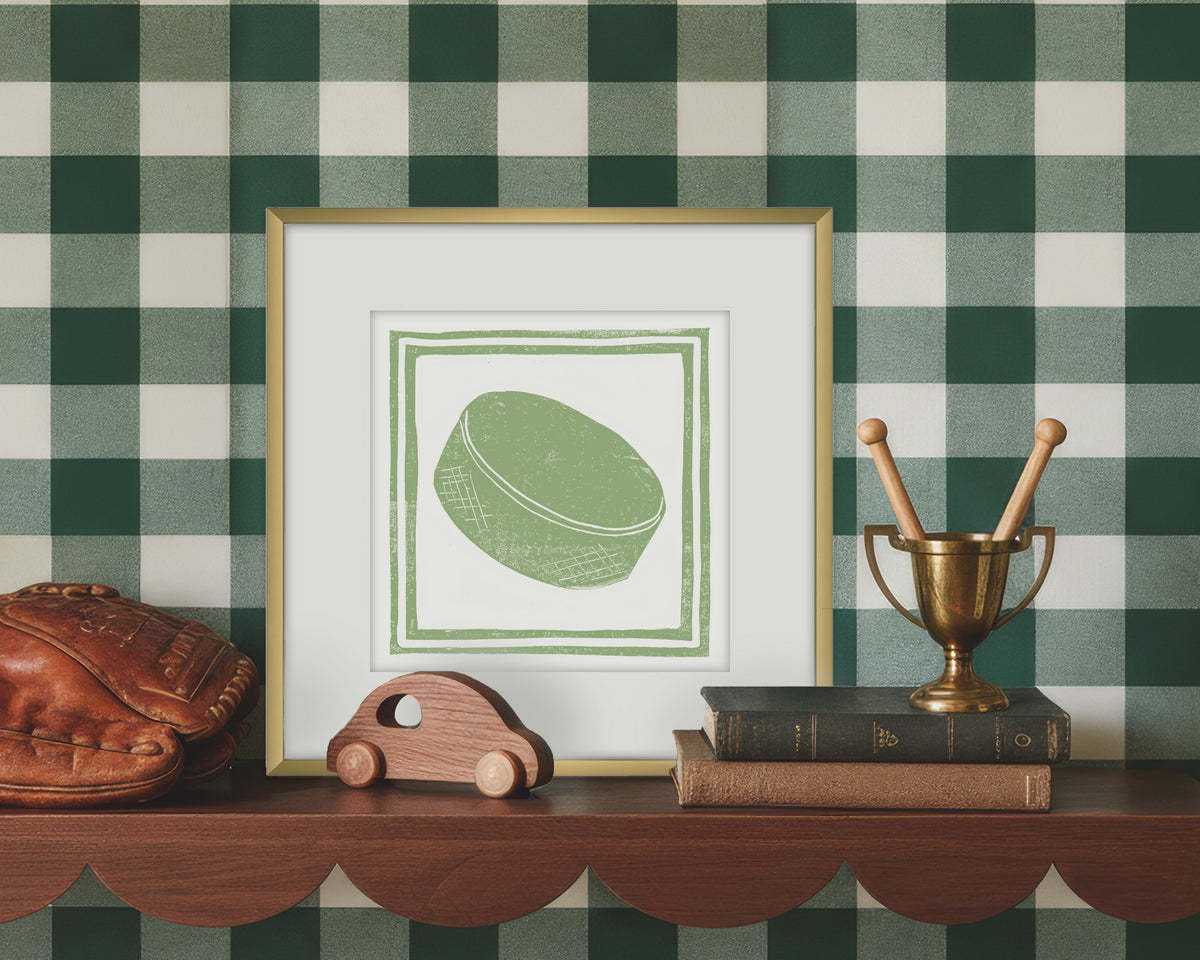 Framed artwork of a green and white hockey puck on a wooden shelf with decorative items against a green checkered wall.