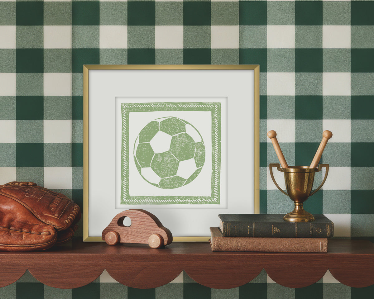 Framed artwork of a green and white soccer ball on a wooden shelf with decorative items against a green checkered wall.