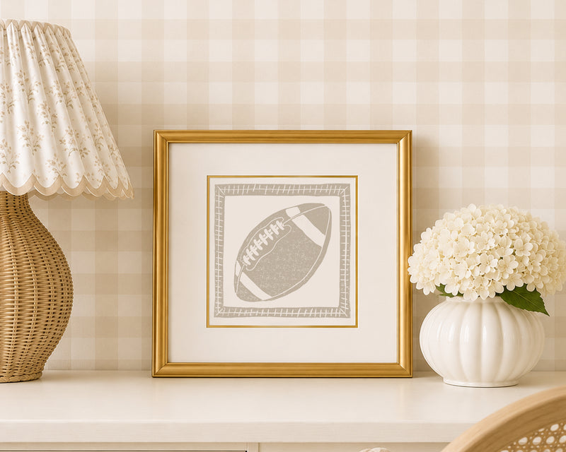 A sand tan and white football art print in a gold matted frame on a styled shelf in a tan and white bedroom