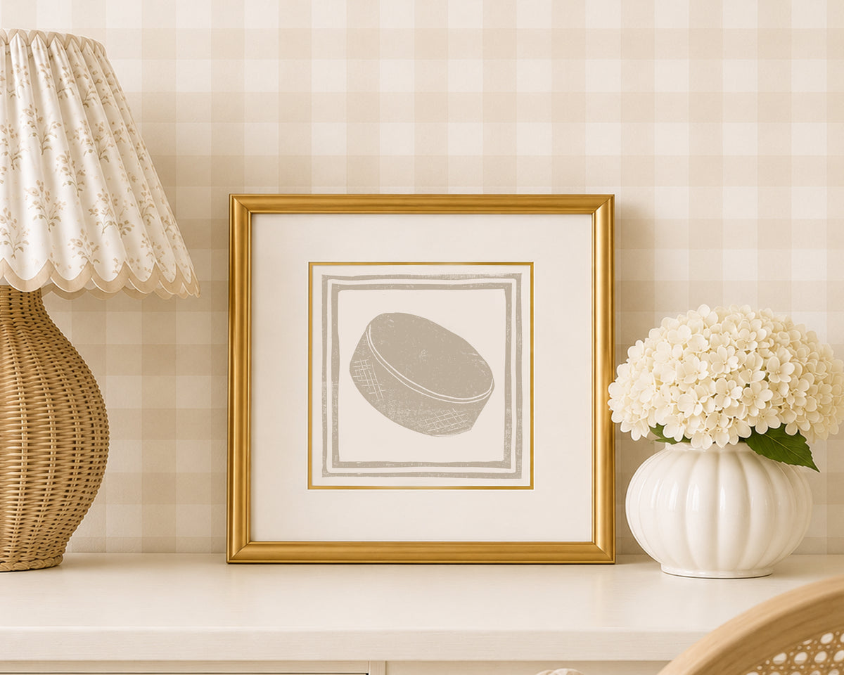 A sand tan and white hockey puck art print in a gold matted frame on a styled shelf in a tan and white bedroom