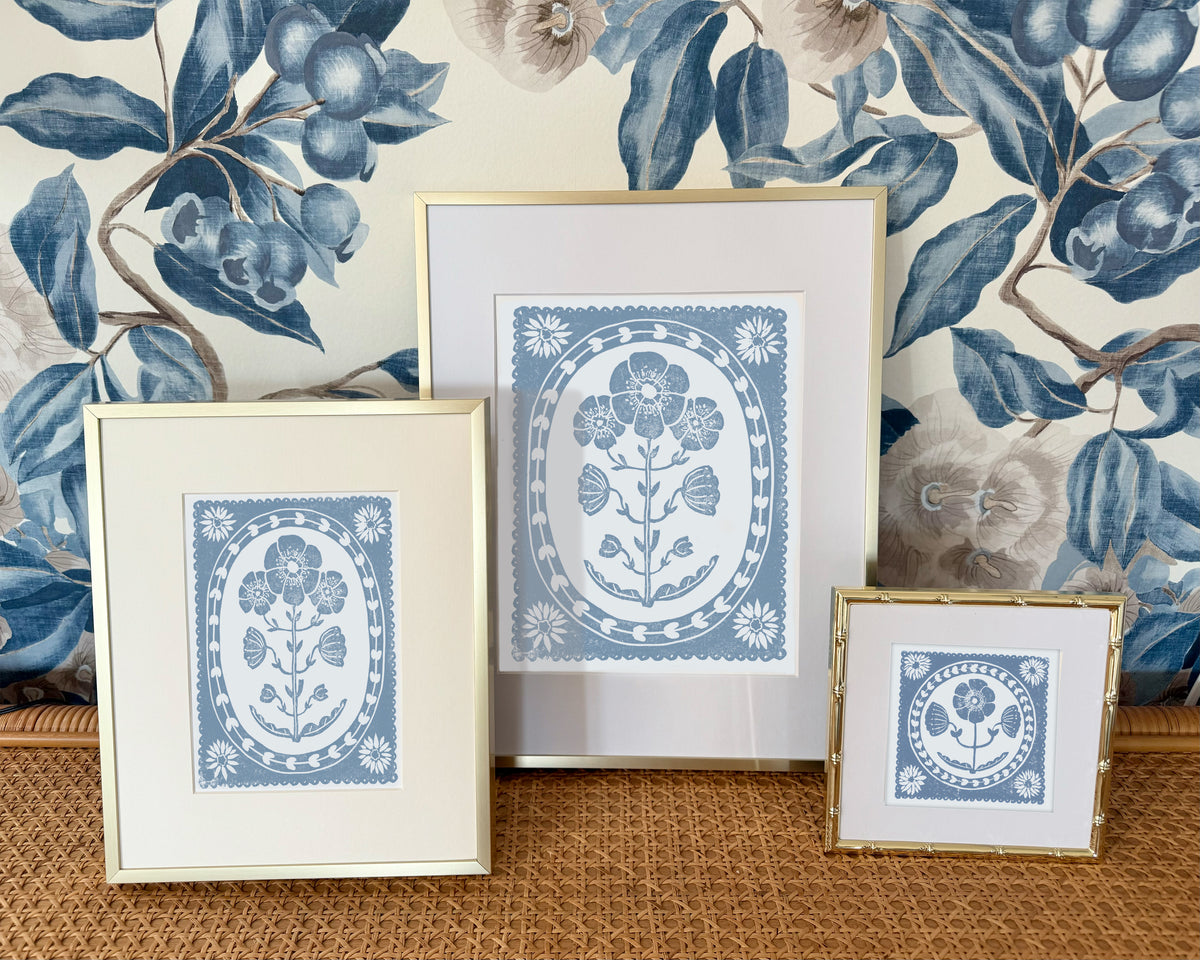 Three framed cornflower blue and white floral prints on a textured surface with decorative wallpaper in the background