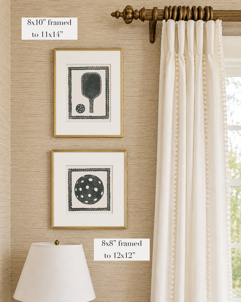 Framed black and white pickleball themed artwork on a tan grasscloth wallpapered wall with a white window treatment and lampshade
