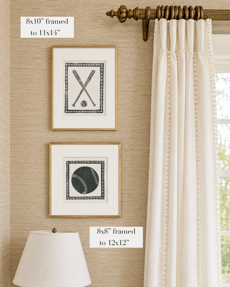 Framed black and white softball themed artwork on a tan grasscloth wallpapered wall with a white window treatment and lampshade