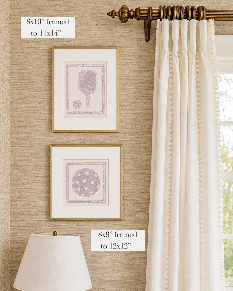 Framed mauve and white pickleball themed artwork on a tan grasscloth wallpapered wall with a white window treatment and lampshade