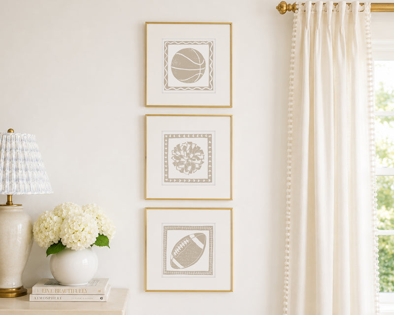Trio of decorative tan colored wall art with sports-themed prints on a white wall, next to a lamp and books.