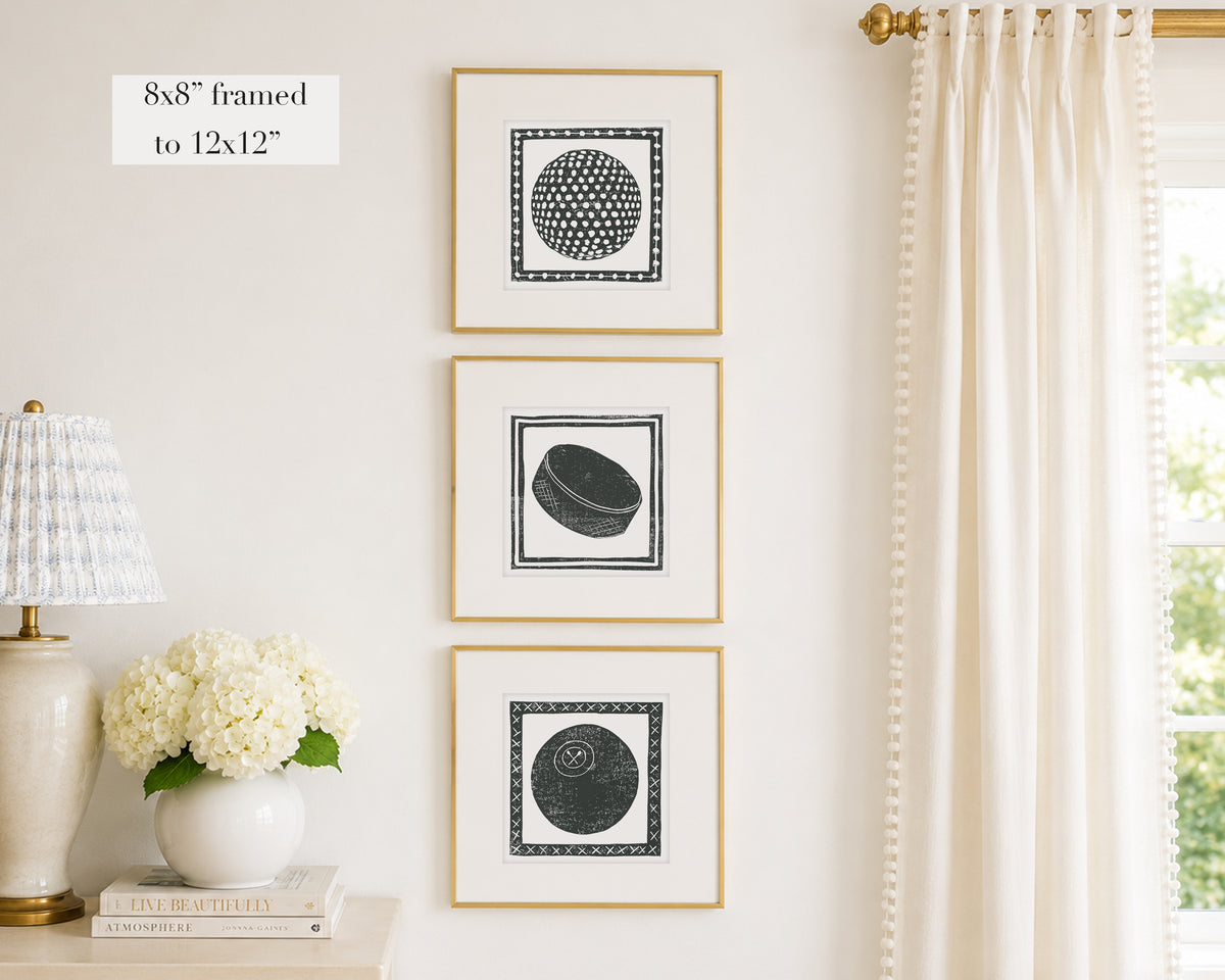 Three framed prints of black and white sport themed block prints on a wall with a lamp and flowers below.