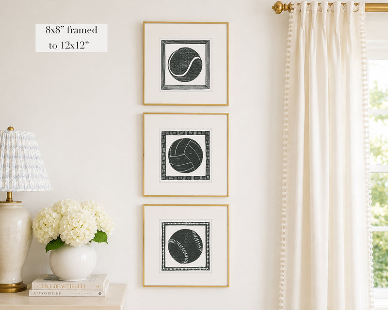 Three framed prints of black and white sport themed block prints on a wall with a lamp and flowers below.