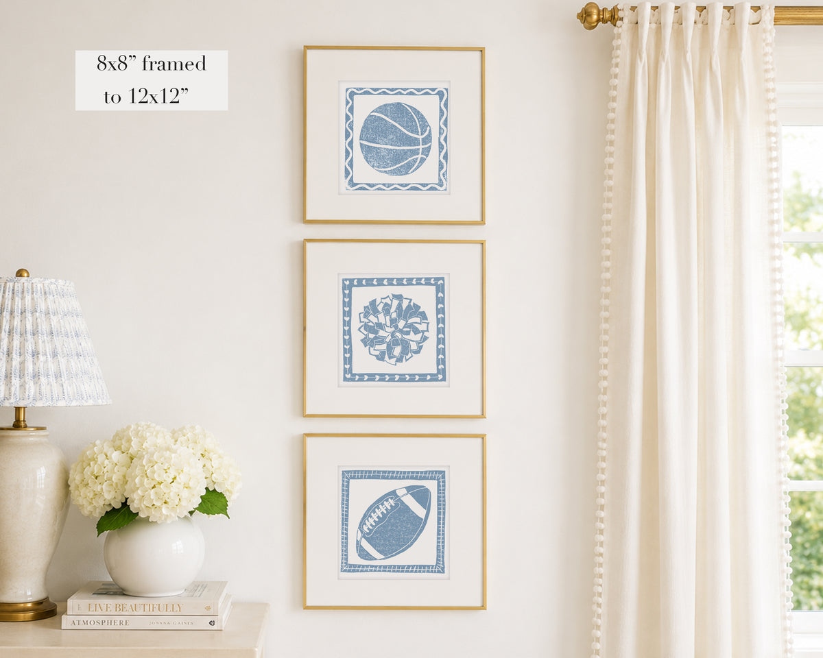 Three framed prints of cornflower blue sport themed block prints on a wall with a lamp and flowers below.