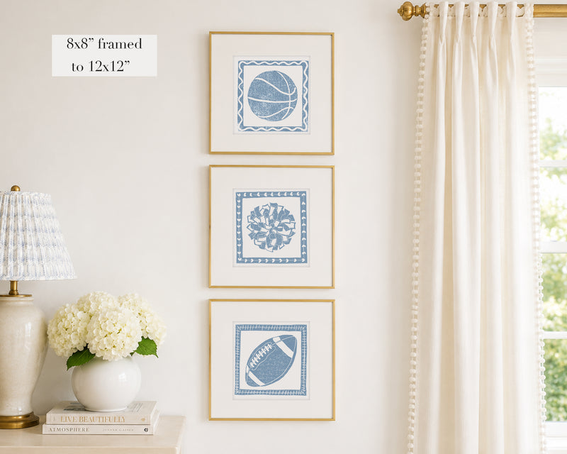 Three framed prints of cornflower blue sport themed block prints on a wall with a lamp and flowers below.