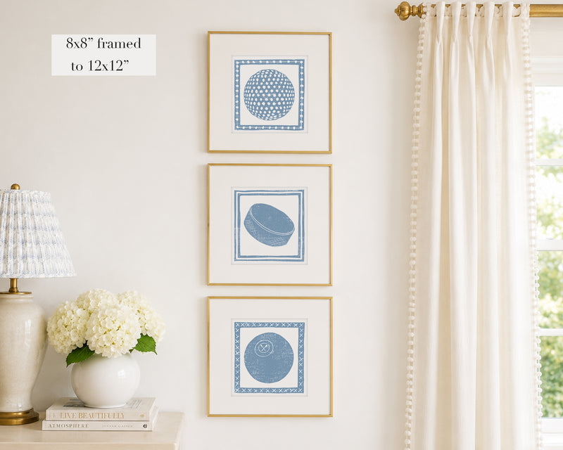 Three framed prints of cornflower blue sport themed block prints on a wall with a lamp and flowers below.