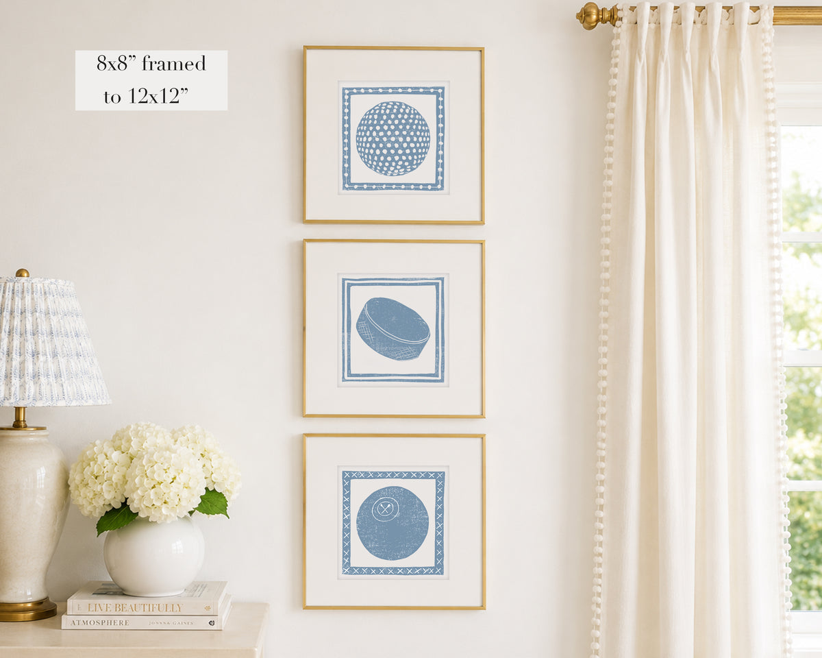 Three framed prints of cornflower blue sport themed block prints on a wall with a lamp and flowers below.