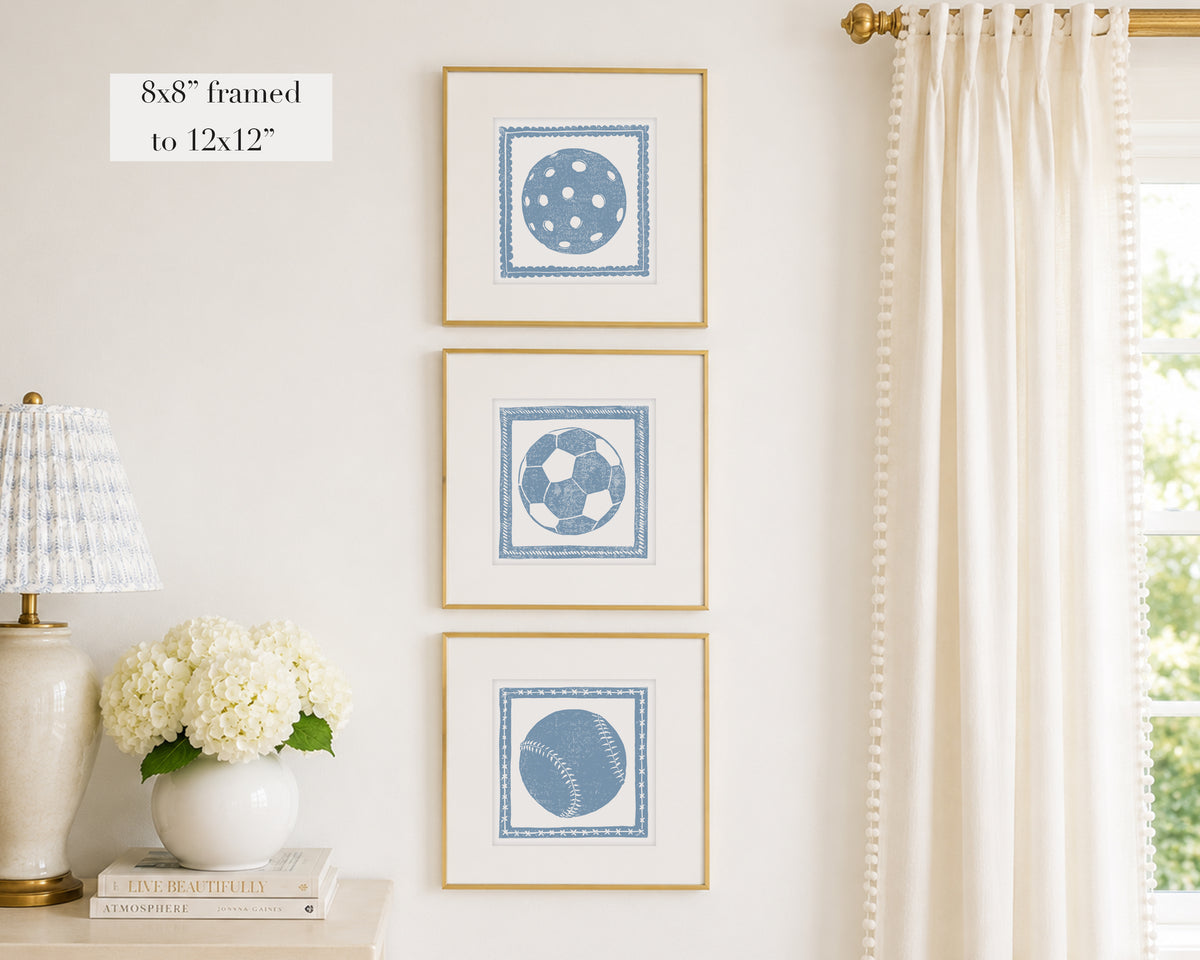 Three framed prints of cornflower blue sport themed block prints on a wall with a lamp and flowers below.