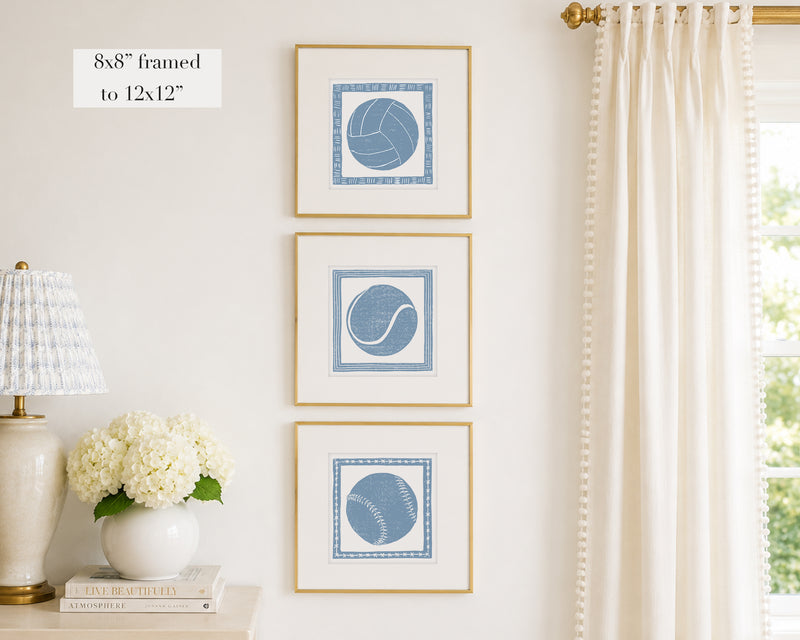 Trio of decorative cornflower blue colored wall art with sports-themed prints on a white wall, next to a lamp and books.