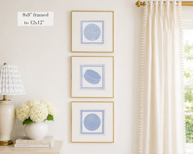 Trio of decorative hydrangea blue colored wall art with sports-themed prints on a white wall, next to a lamp and books.