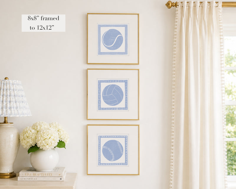 Trio of decorative hydrangea blue colored wall art with sports-themed prints on a white wall, next to a lamp and books.