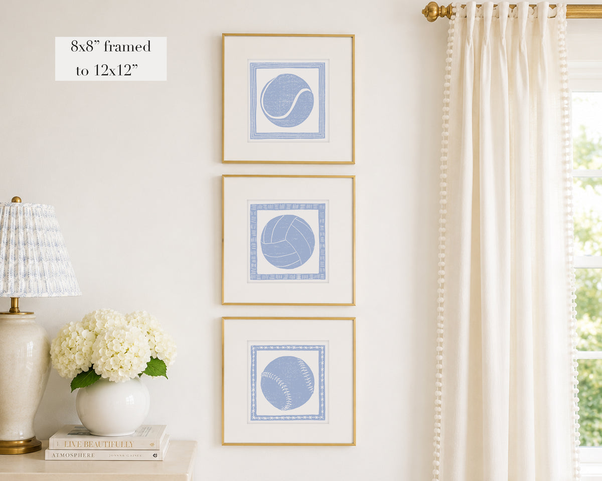 Trio of decorative hydrangea blue colored wall art with sports-themed prints on a white wall, next to a lamp and books.