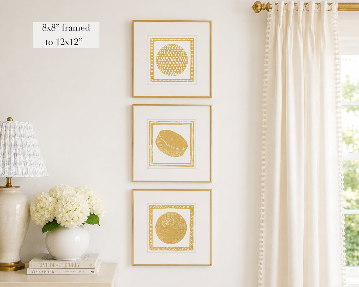 Trio of decorative mustard yellow colored wall art with sports-themed prints on a white wall, next to a lamp and books.