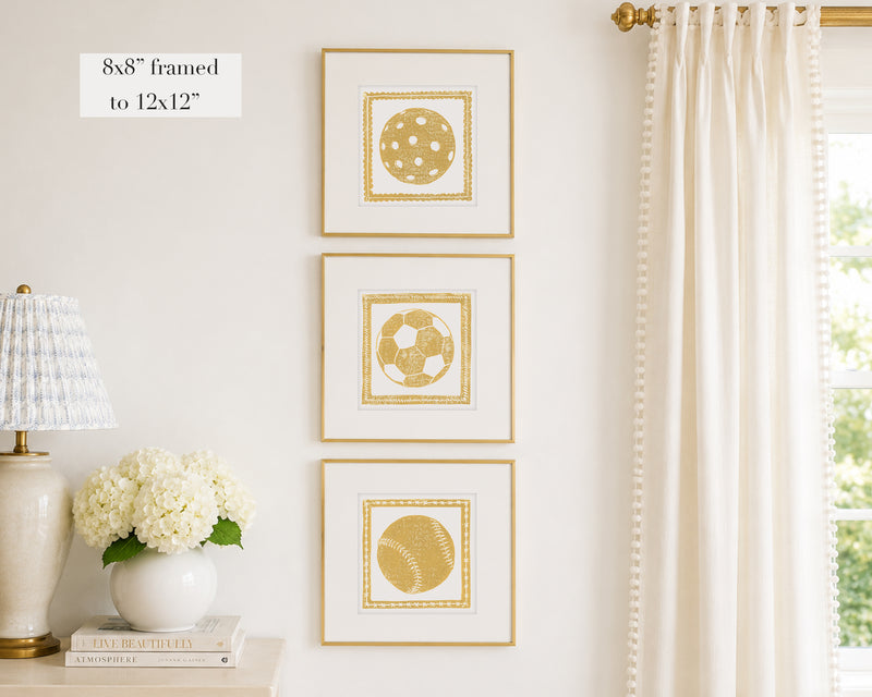 Trio of decorative mustard yellow colored wall art with sports-themed prints on a white wall, next to a lamp and books.
