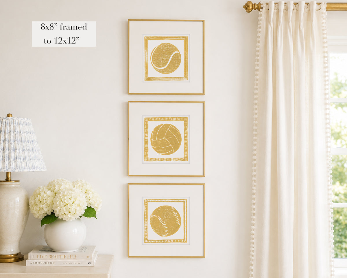 Trio of decorative mustard yellow colored wall art with sports-themed prints on a white wall, next to a lamp and books.