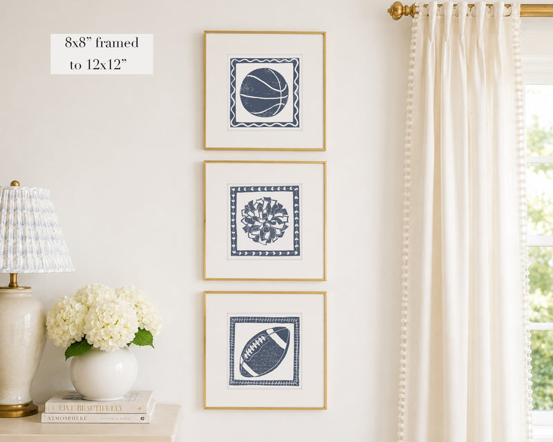 Trio of decorative navy blue colored wall art with sports-themed prints on a white wall, next to a lamp and books.