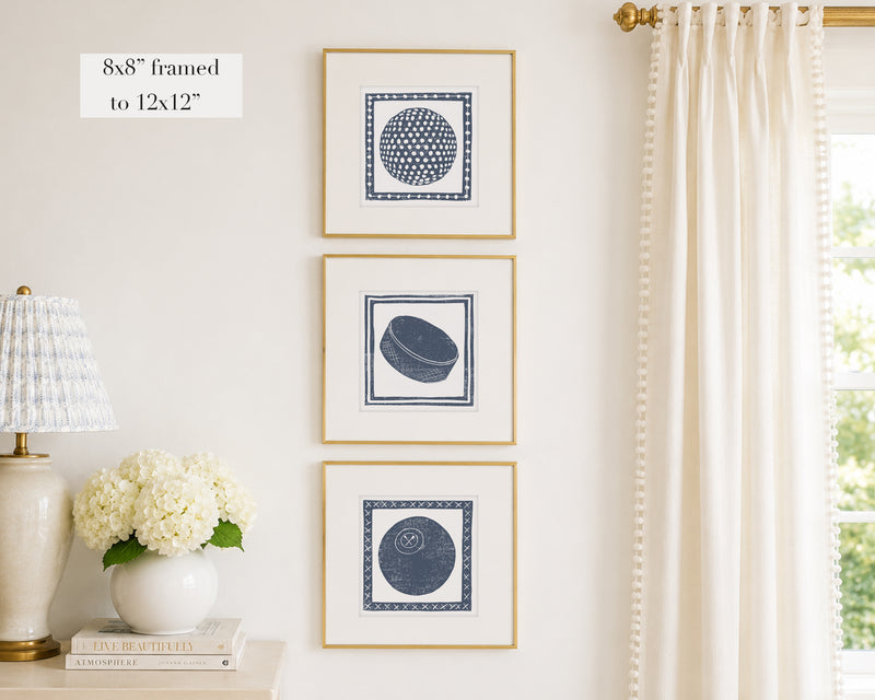 Trio of decorative navy blue colored wall art with sports-themed prints on a white wall, next to a lamp and books.