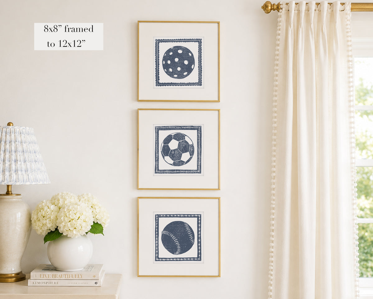 Trio of decorative navy blue colored wall art with sports-themed prints on a white wall, next to a lamp and books.
