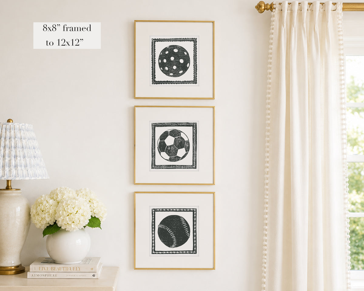 Three framed prints of black and white sport themed block prints on a wall with a lamp and flowers below.