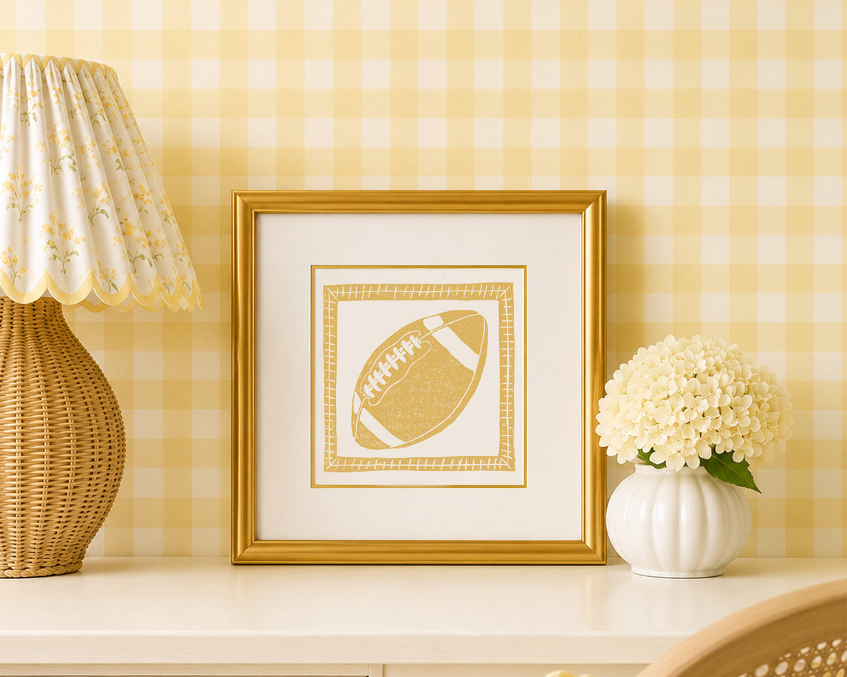 A mustard yellow and white football art print in a gold matted frame on a styled shelf in a yellow and white bedroom