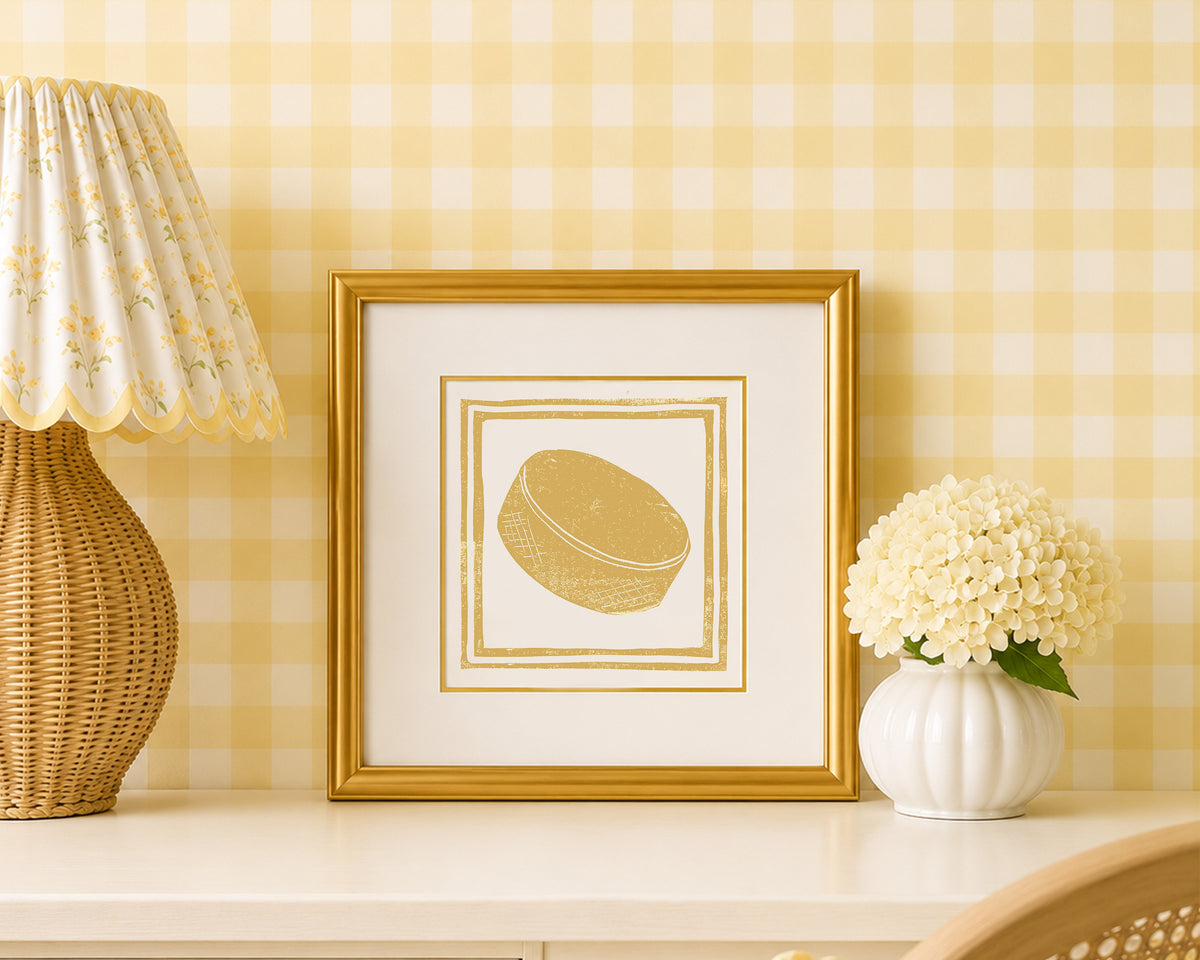 A mustard yellow and white hockey puck art print in a gold matted frame on a styled shelf in a yellow and white bedroom