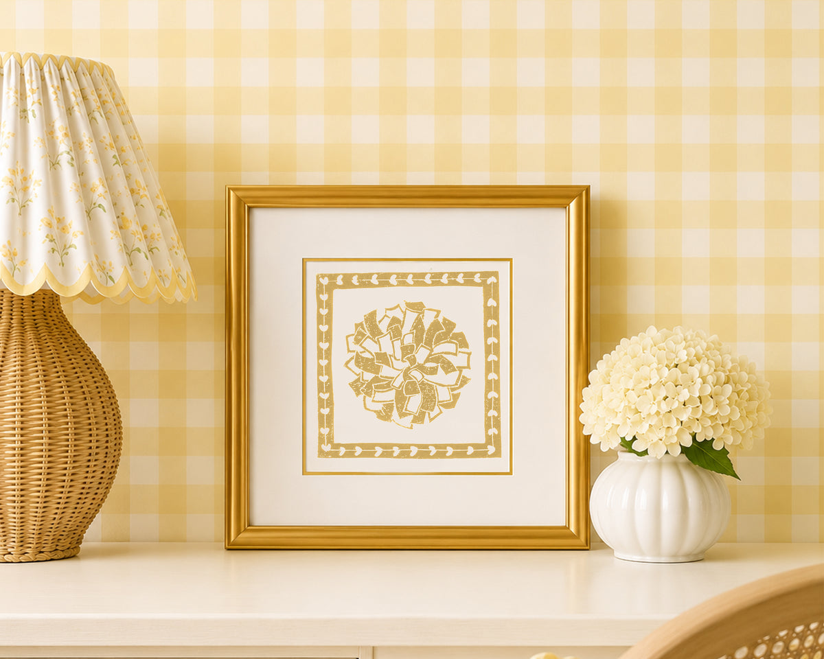 A mustard yellow and white pom pon art print in a gold matted frame on a styled shelf in a yellow and white bedroom