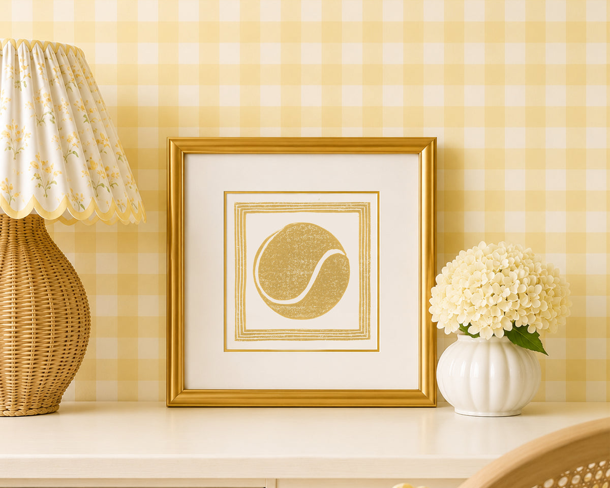 A mustard yellow and white tennis ball art print in a gold matted frame on a styled shelf in a yellow and white bedroom