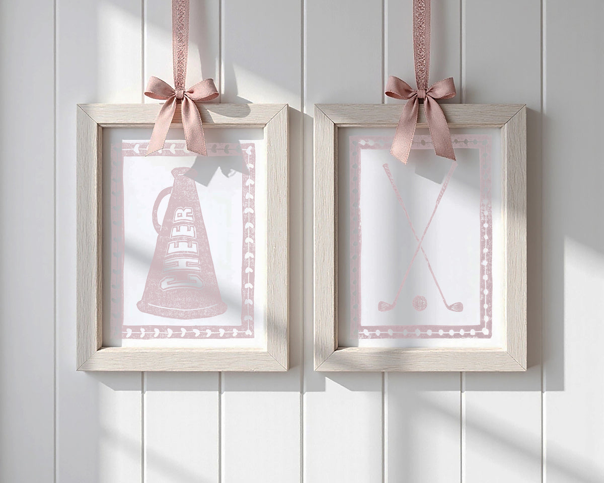 Two framed pictures with golf clubs and ball and cheer megaphone, each hanging on a pink ribbon against a white paneled wall.