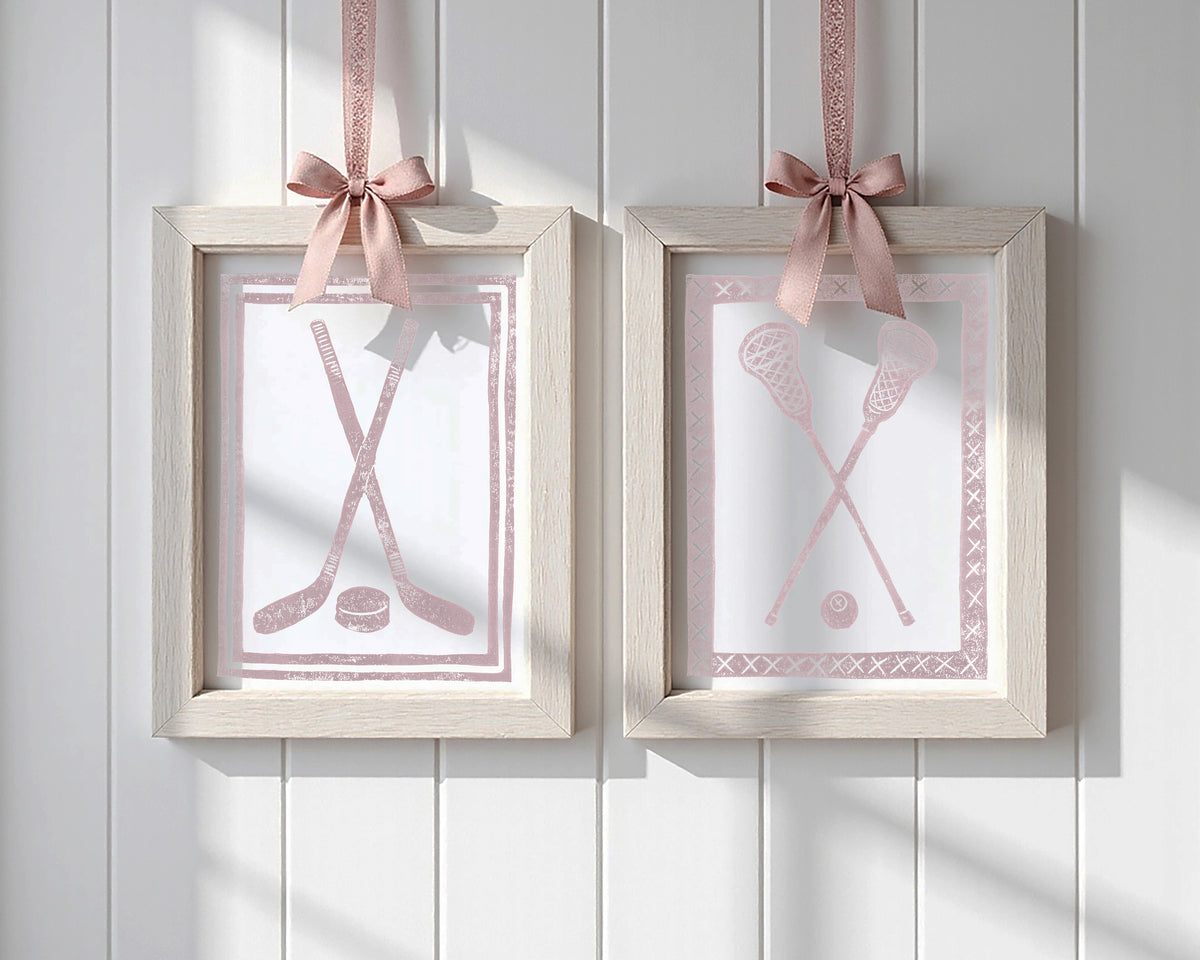 Two framed pictures with hockey sticks and lacrosse crosses and balls, each hanging on a pink ribbon against a white paneled wall.