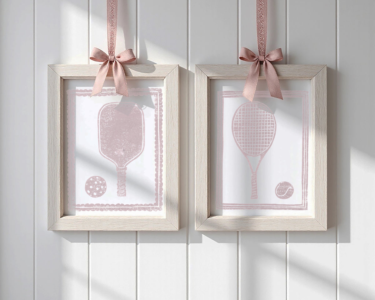 Two framed pictures with pickleball, and tennis rackets and balls, each hanging on a pink ribbon against a white paneled wall.