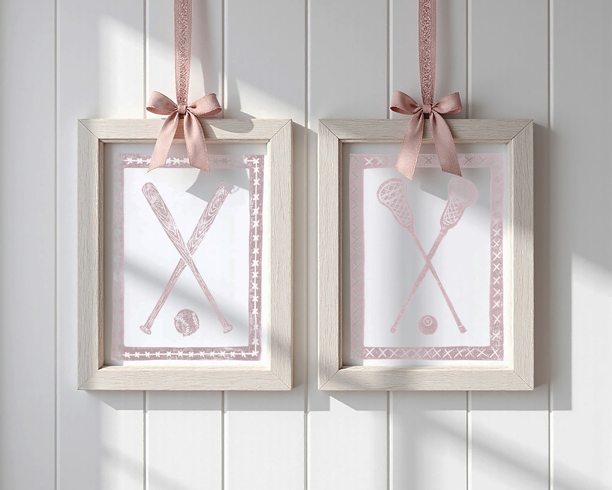 Two framed pictures with baseball bats and lacrosse sticks and balls, each hanging on a pink ribbon against a white paneled wall.